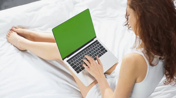 Hochwinkelaufnahme der lockigen jungen Frau, die auf dem Bett sitzt und Laptop mit grünem Bildschirm benutzt — Stockfoto
