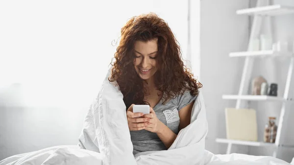 Happy young woman wrapped in white blanket texting on smartphone in bedroom — Stock Photo