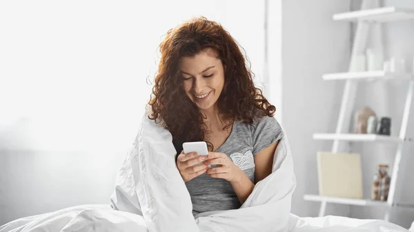 Щаслива молода жінка, загорнута в біле ковдру повідомлення на смартфон в спальні — стокове фото