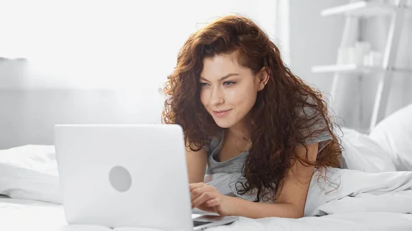 Joven freelancer satisfecho usando el ordenador portátil en la cama - foto de stock