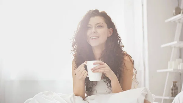 Glückliche und lockige Frau mit einer Tasse Kaffee im Bett — Stockfoto