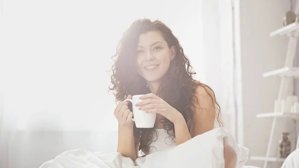 Cheerful curly woman holding cup of coffee in bed — Stock Photo