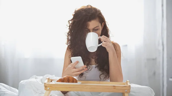 Mujer rizada beber café y el uso de teléfono inteligente cerca de la bandeja con croissant en la cama - foto de stock