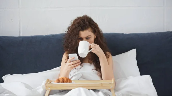 Молодая кудрявая женщина пьет кофе и держит смартфон рядом с подносом с круассаном на кровати — стоковое фото