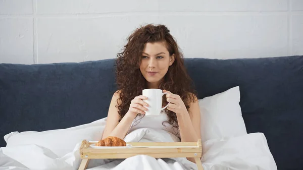 Улыбающаяся женщина держит чашку кофе возле подноса с круассаном на кровати — стоковое фото