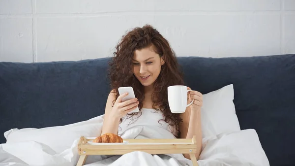 Sorrindo e encaracolado mulher segurando xícara de café e usando smartphone perto bandeja com croissant saboroso na cama — Fotografia de Stock