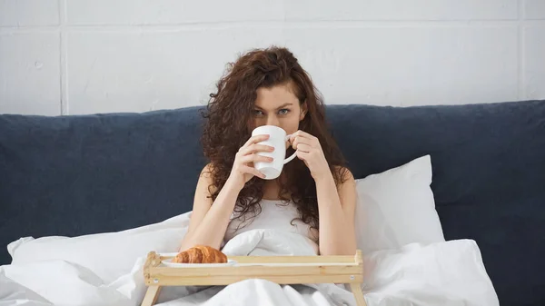 Mulher encaracolado beber café perto da bandeja com croissant na cama — Fotografia de Stock