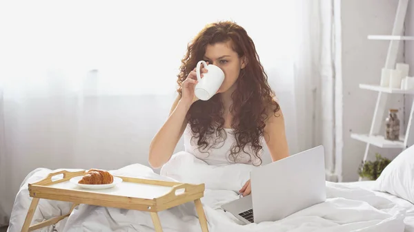 Lockige junge Frau trinkt Kaffee in der Nähe Tablett mit leckeren Croissant und Laptop auf dem Bett — Stockfoto