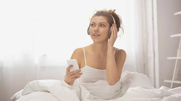 Mulher acordada em fones de ouvido sem fio ouvindo música e segurando smartphone no quarto — Fotografia de Stock