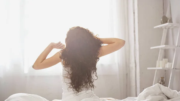 Rückansicht einer jungen lockigen Frau, die sich dehnt, während sie auf dem Bett sitzt — Stockfoto