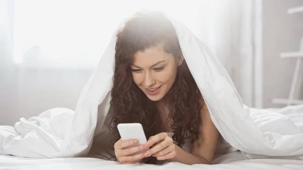 Mujer joven y feliz usando el teléfono inteligente mientras está acostado debajo de la manta - foto de stock