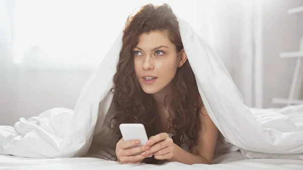Pensive young woman using smartphone while lying under blanket — Stock Photo