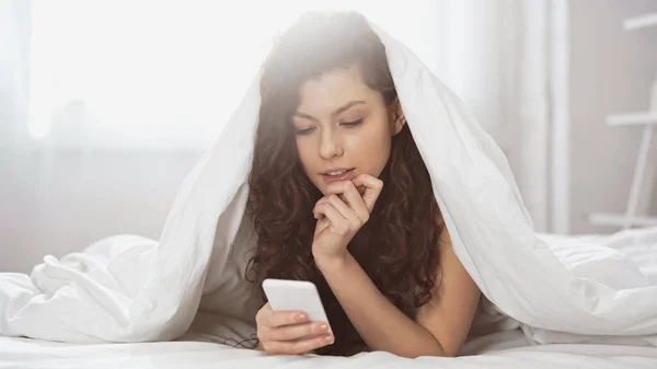 Nachdenkliche junge Frau benutzt Smartphone, während sie unter Decke liegt — Stockfoto