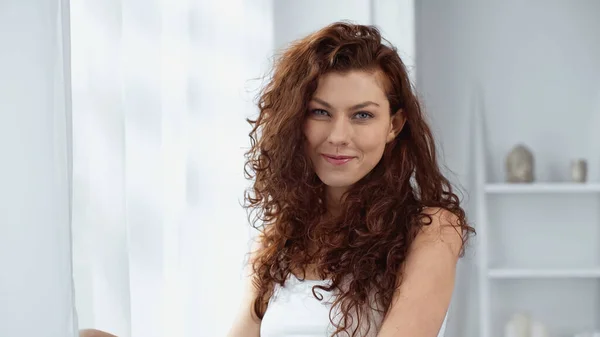 Curly woman smiling while looking at camera near curtain at home — Stock Photo