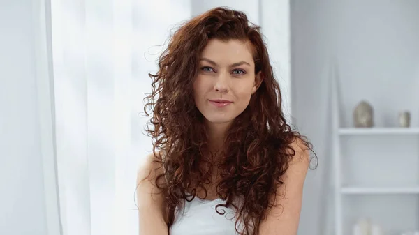 Young and curly woman smiling while looking at camera near curtain at home — Stock Photo