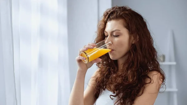 Curly young woman holding glass and drinking orange juice at home — Stock Photo