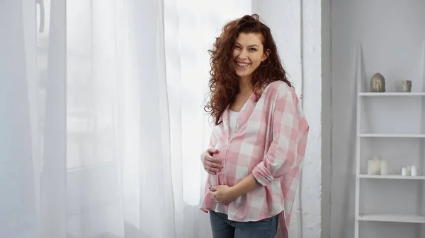 Pregnant and curly woman smiling near white curtain — Stock Photo