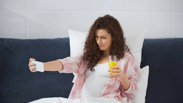 Кудрявая и беременная женщина делает селфи, держа стакан апельсинового сока в спальне — стоковое фото