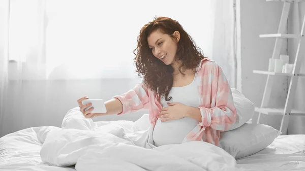 Smiling and pregnant woman taking selfie in bedroom — Stock Photo