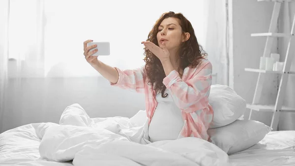 Lockige und schwangere Frau verschickt Luftkuss, während sie ein Selfie im Schlafzimmer macht — Stockfoto