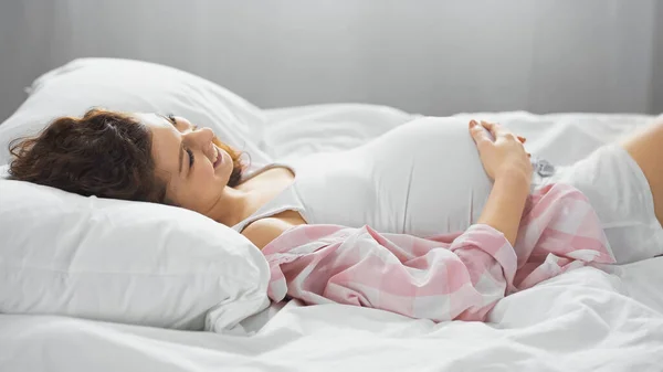 Feliz joven tocando el vientre mientras está acostado en la cama - foto de stock