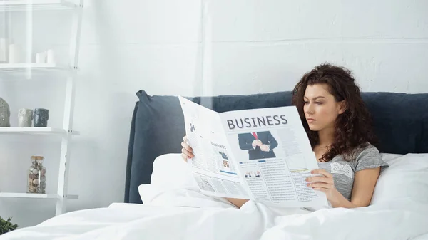 Mujer joven y rizada leyendo periódico de negocios en la cama - foto de stock