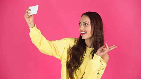 Smiling young adult woman taking selfie on cellphone with peace gesture isolated on pink — Stock Photo