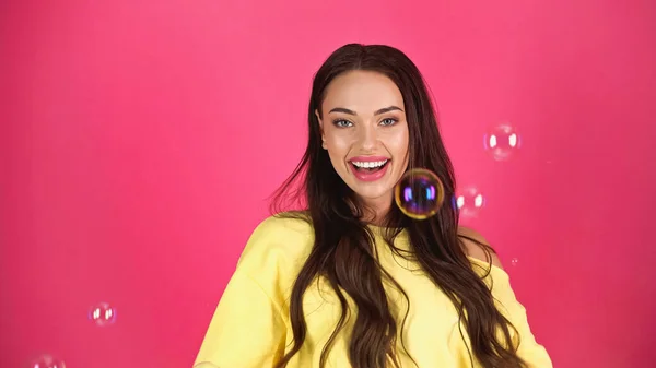 Positive junge erwachsene Frau in gelber Bluse mit Seifenblasen auf rosa Hintergrund — Stockfoto