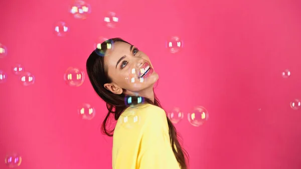 Happy young adult woman in yellow blouse with soap bubbles on pink background - foto de stock