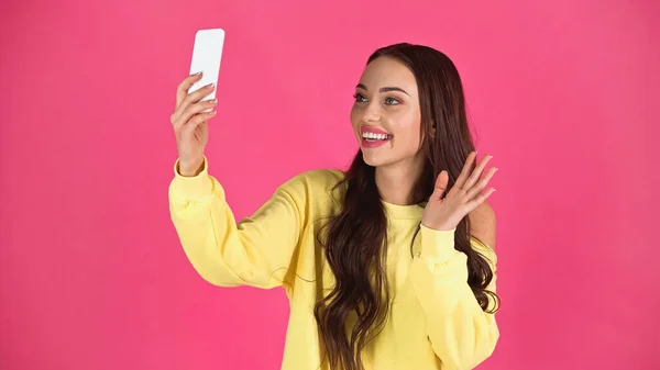 Smiling young adult woman having video call and waving hand to cellphone isolated on pink - foto de stock