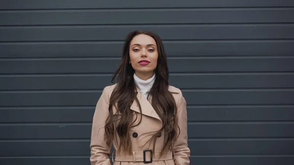 Confident woman in beige trench coat and turtleneck looking at camera on textured grey background — Photo de stock