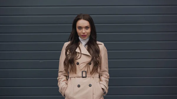 Attractive young adult woman in beige trench coat and turtleneck looking at camera on textured grey background — Fotografia de Stock