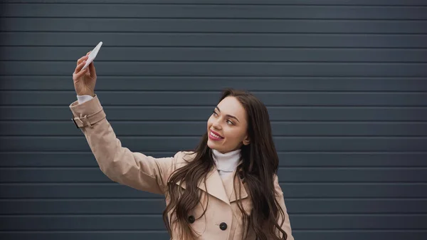 Усміхнена молода доросла жінка бере селфі з мобільним телефоном на текстурованому сірому фоні — стокове фото