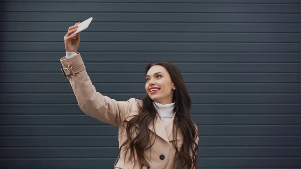Усміхнена молода доросла жінка в бежевому пальто бере селфі з мобільним телефоном на сірому текстурованому фоні — стокове фото