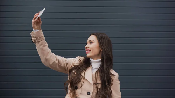 Hübsche junge erwachsene Frau macht Selfie mit Handy auf grau strukturiertem Hintergrund — Stockfoto
