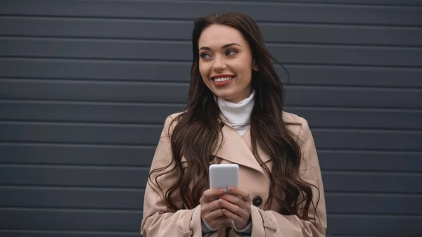 Smiling young adult woman holding cellphone in hands on grey textured background — Photo de stock