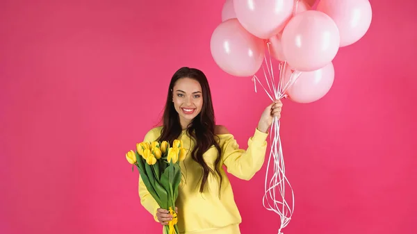 Усміхнена молода доросла жінка тримає букет тюльпанів і повітряних кульок ізольовано на рожевому — стокове фото
