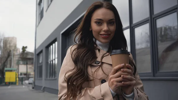 Усміхнена молода доросла жінка тримає паперову чашку біля будівлі на вулиці — стокове фото