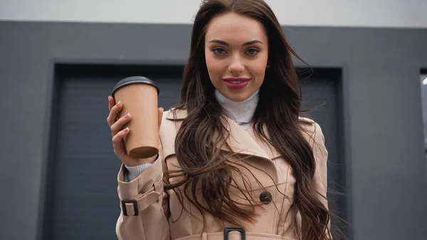 Positive young adult woman in autumn outfit holding paper cup and looking at camera on urban background — Stock Photo