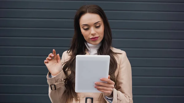 Junge erwachsene Frau im beigen Trenchcoat mit Tablette auf strukturiertem grauen Hintergrund — Stockfoto