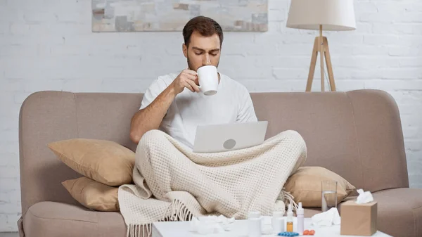 Kranker Mann sitzt auf Sofa mit Laptop und trinkt Tee am Kaffeetisch mit Tabletten in Flaschen — Stockfoto