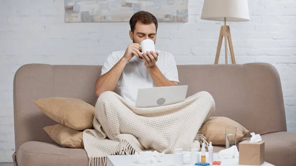 Sick freelancer sitting on sofa with laptop and drinking tea near coffee table with pills in bottles — Photo de stock