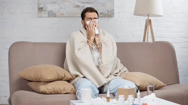 Kranker Mann sitzt auf Sofa und niest mit Getränken und Flaschen in Taschentuch neben Couchtisch — Stockfoto