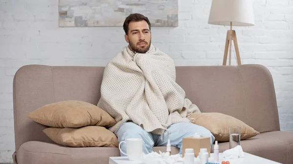 Sick man wrapped in blanket sitting on sofa in living room — Photo de stock