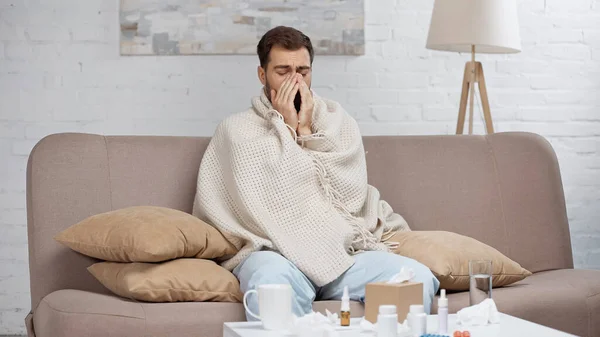 Sick man sneezing near coffee table with drinks and bottles — Stock Photo