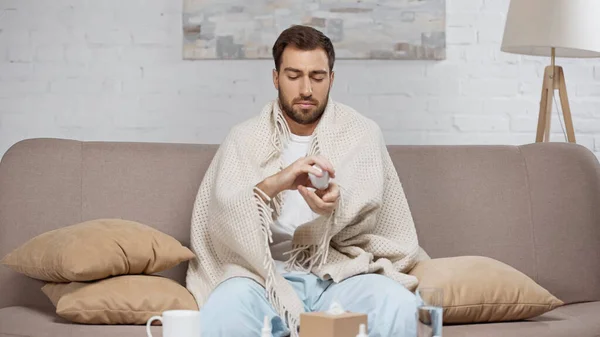 Kranker Mann sitzt auf Sofa und hält Flasche mit Tabletten neben Kaffeetisch mit Getränken — Stockfoto