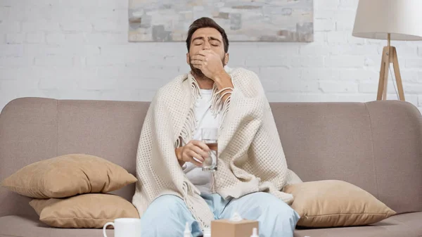 Sick man sitting on sofa and taking pill near coffee table with cup and bottles — Stock Photo