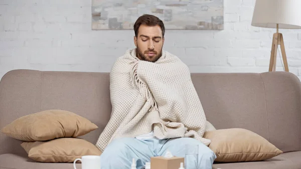 Ill man wrapped in blanket sitting on sofa in living room - foto de stock