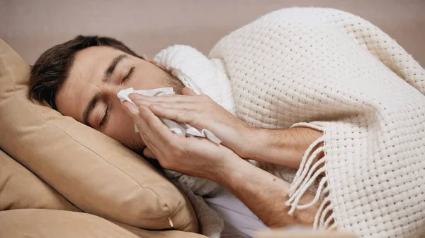 Sick man lying under blanket on sofa and sneezing in tissue - foto de stock