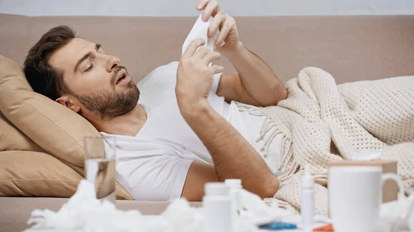 Kranker Mann liegt unter Decke und niest in der Nähe von Medikamenten und zerknittertem Gewebe — Stockfoto